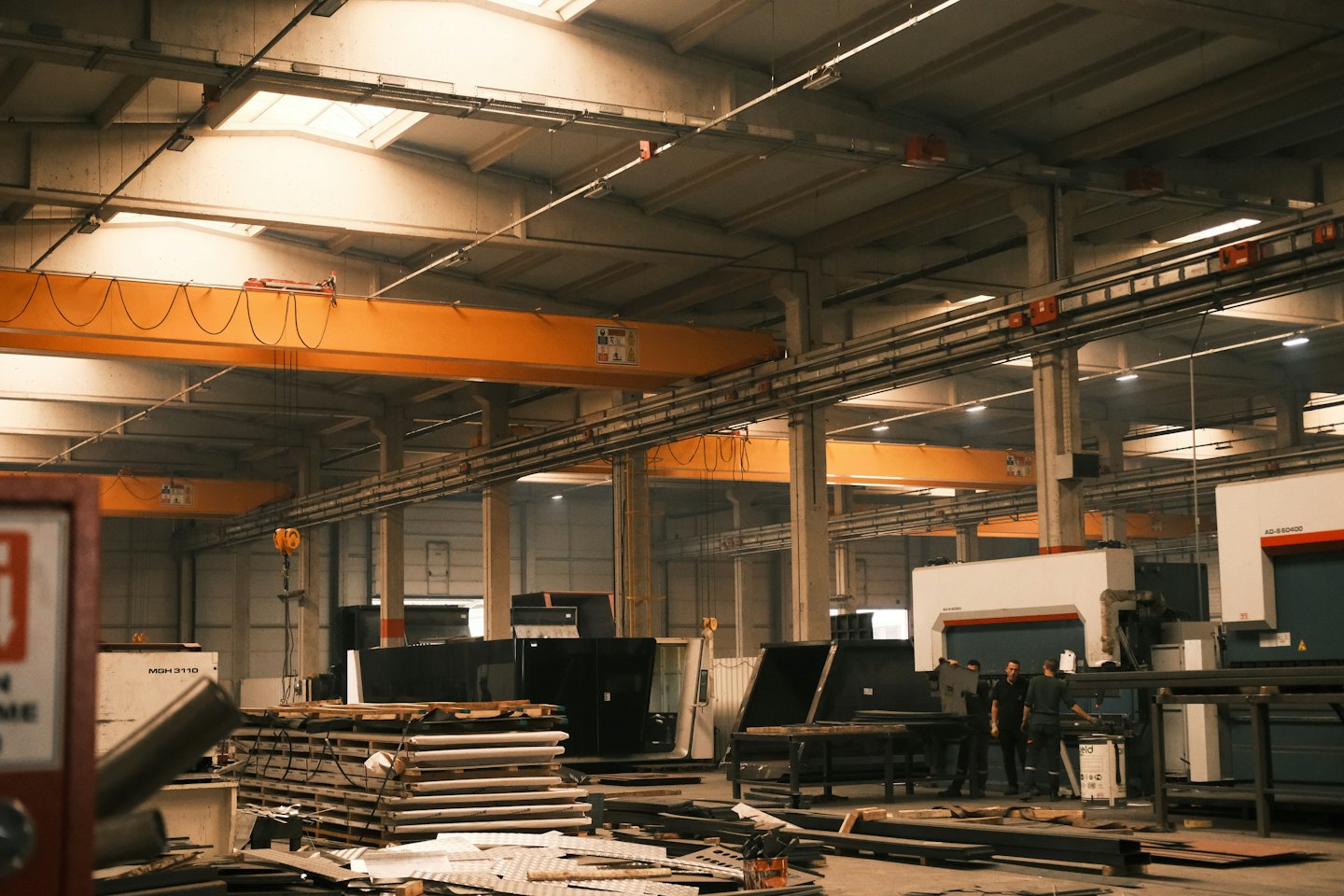 Workers operate sheet metal fabrication equipment under orange cranes.